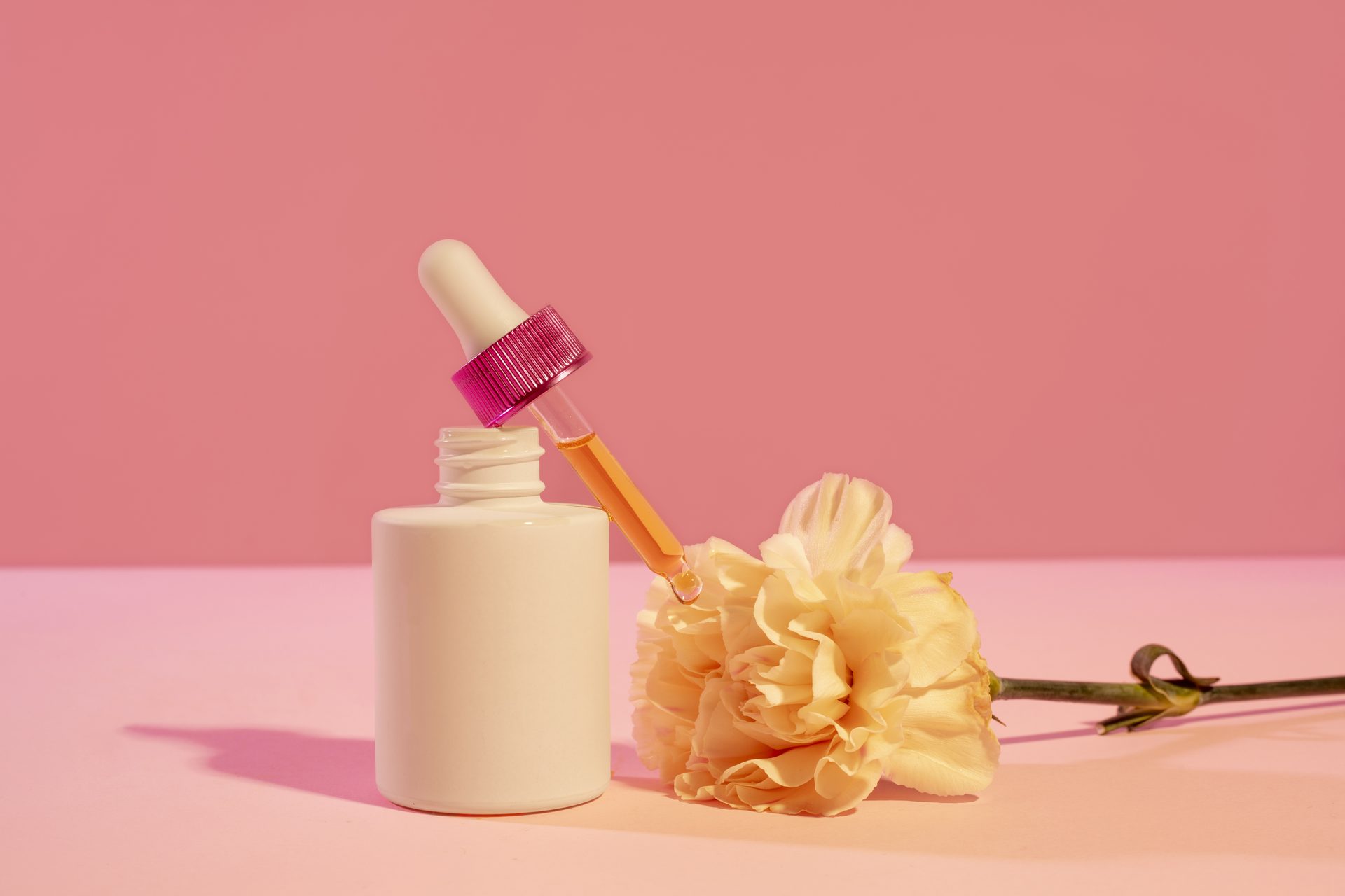 Frasco cuentagotas blanco con tapa rosa junto a un clavel amarillo pálido sobre fondo rosa
