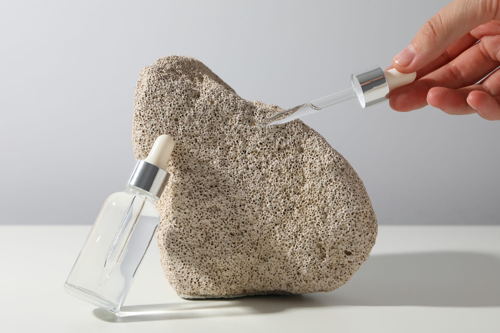 Hand holding a glass dropper over a porous beige stone, with a clear serum bottle leaning against it
