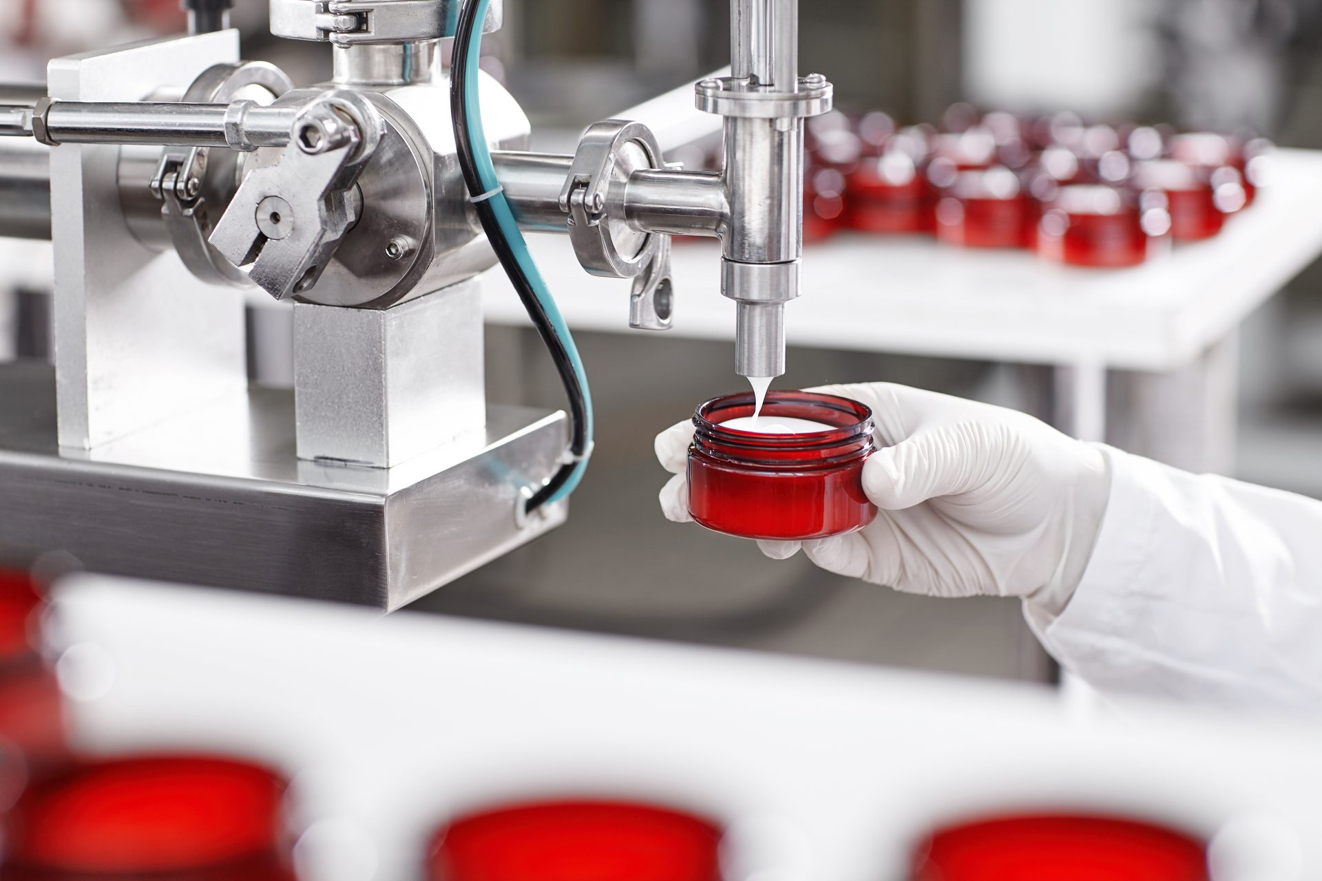Gloved hand holding a red jar under a stainless-steel filling nozzle dispensing white cream