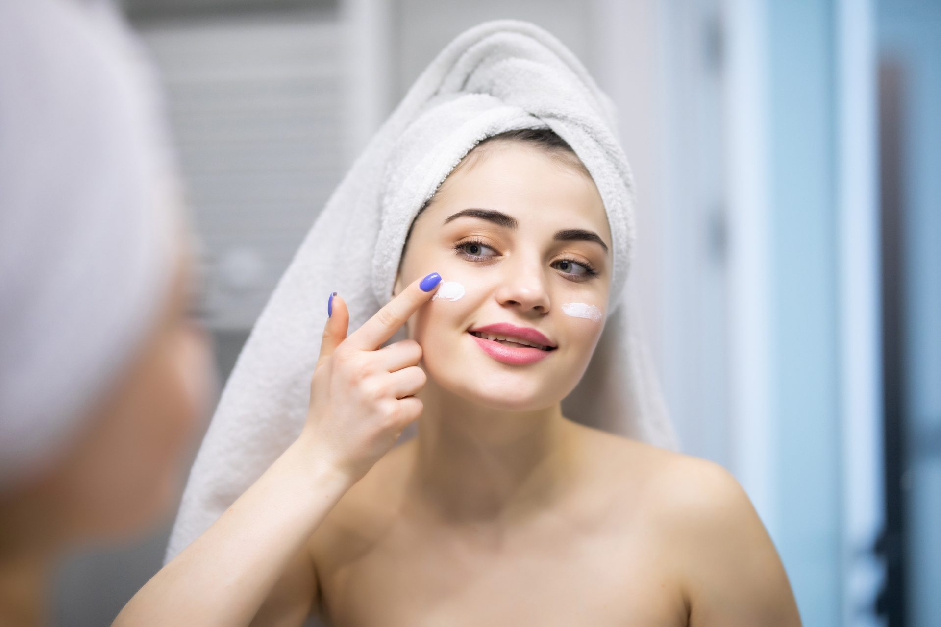 Smiling woman in a towel turban applying dabs of white cream to her cheek in a mirror