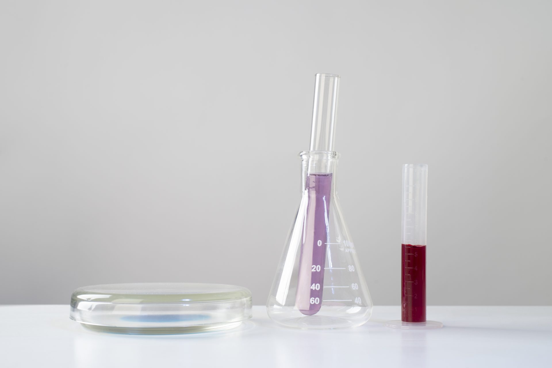 Petri dish, conical flask holding a violet-filled tube, and a cylinder of dark red liquid on white