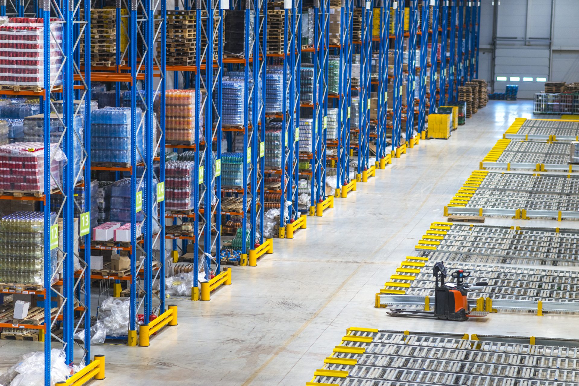 Gran almacén con altas estanterías azules para palés, transportadores de rodillos y una transpaleta naranja