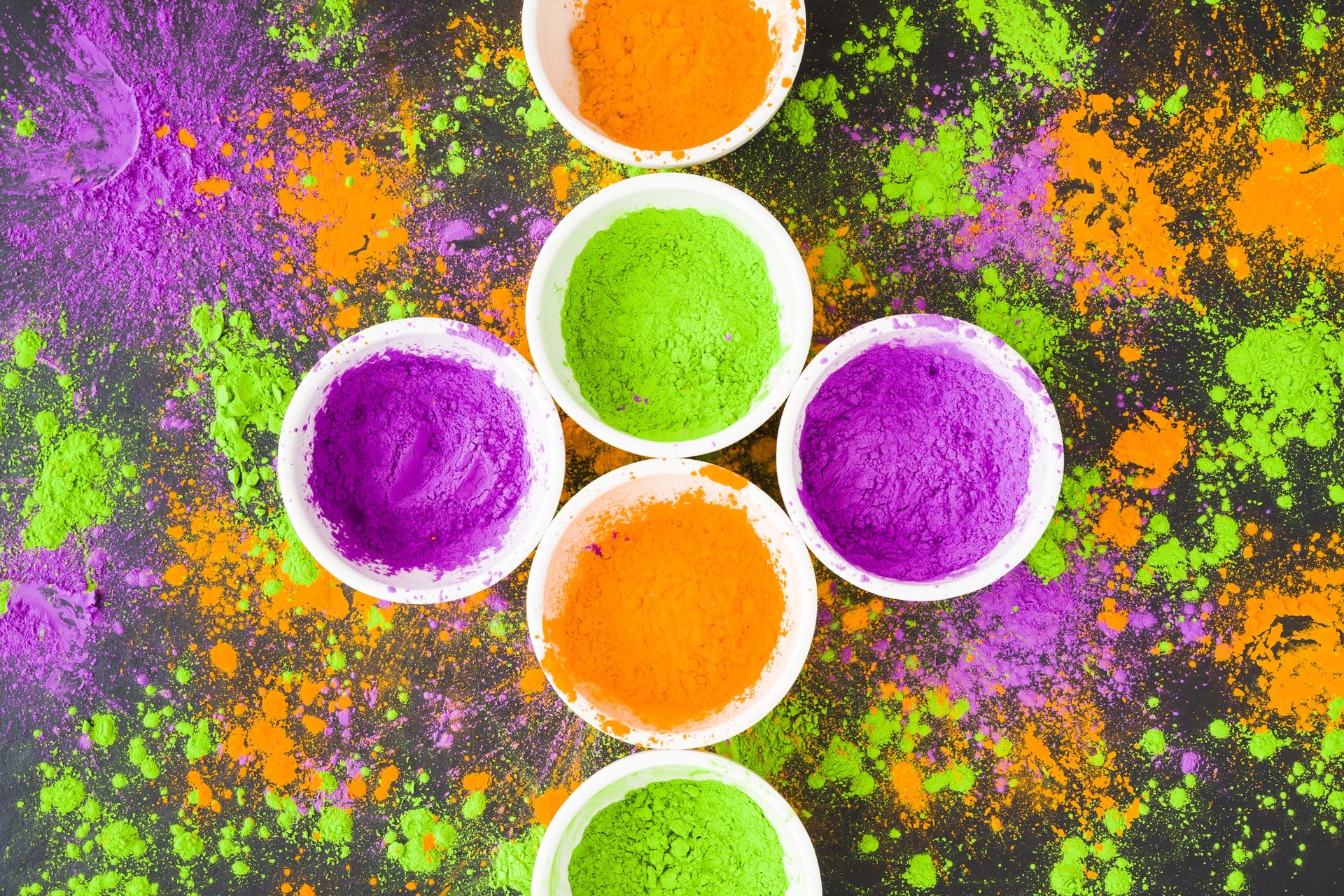 Cuencos blancos con pigmento en polvo naranja, verde y morado entre color esparcido sobre superficie oscura