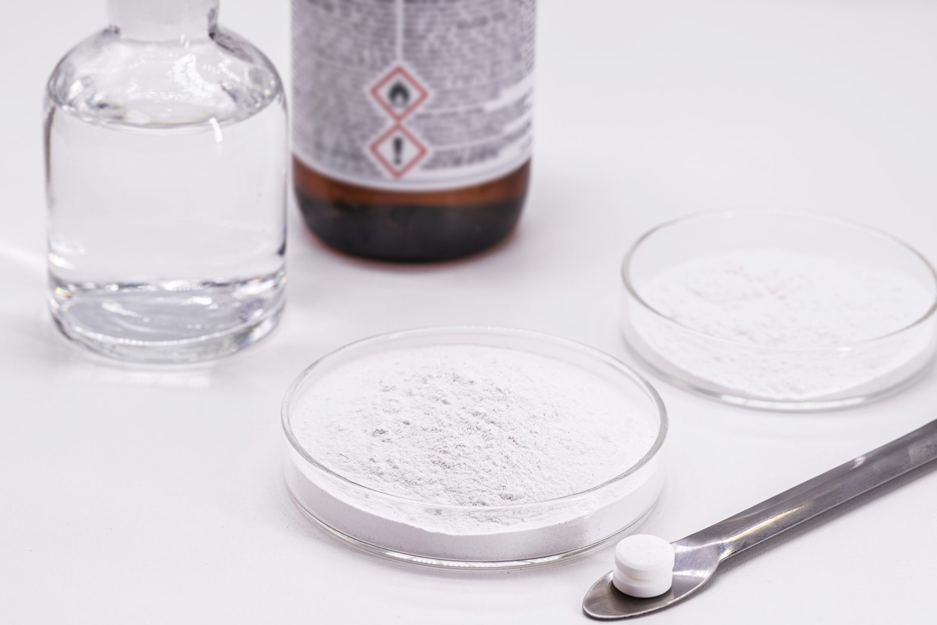White powder in glass petri dishes beside a clear bottle, a brown bottle with hazard symbols and a spatula