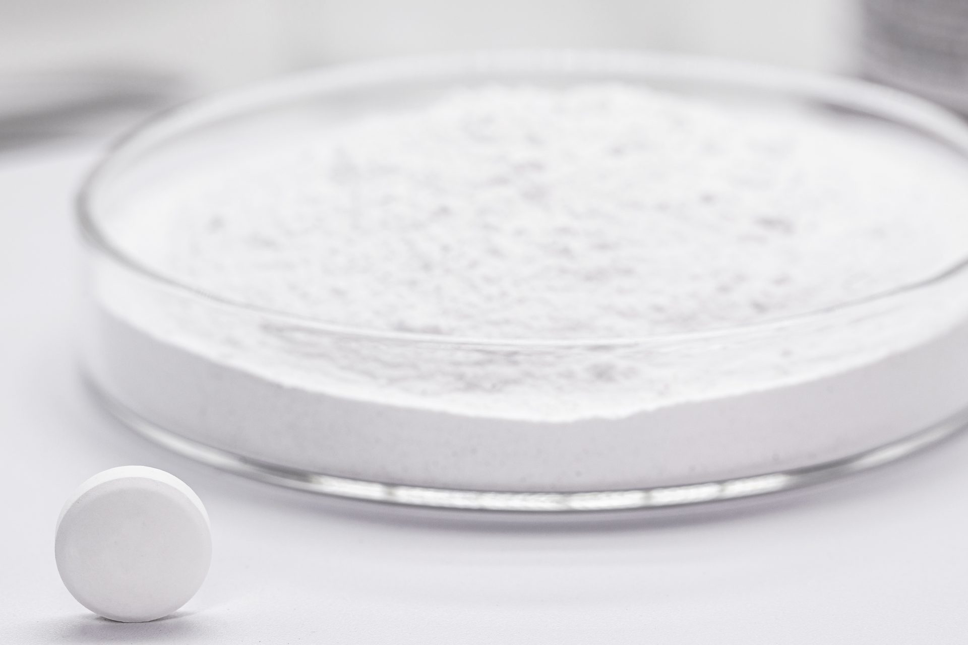Recipiente de vidrio lleno de polvo blanco junto a una pastilla blanca sobre una superficie blanca