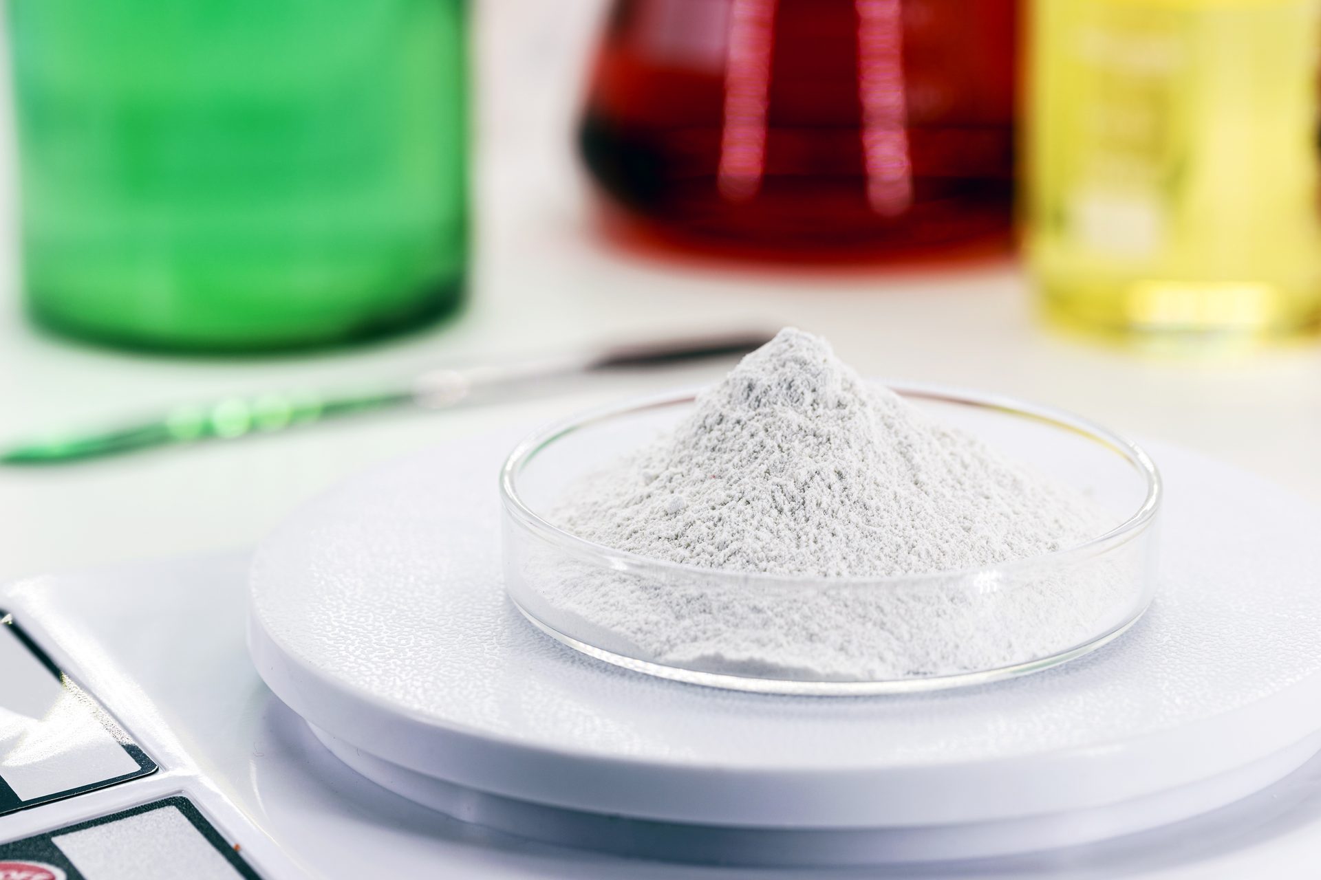 Petri dish of white powder on a laboratory balance, with green, red and yellow liquids behind