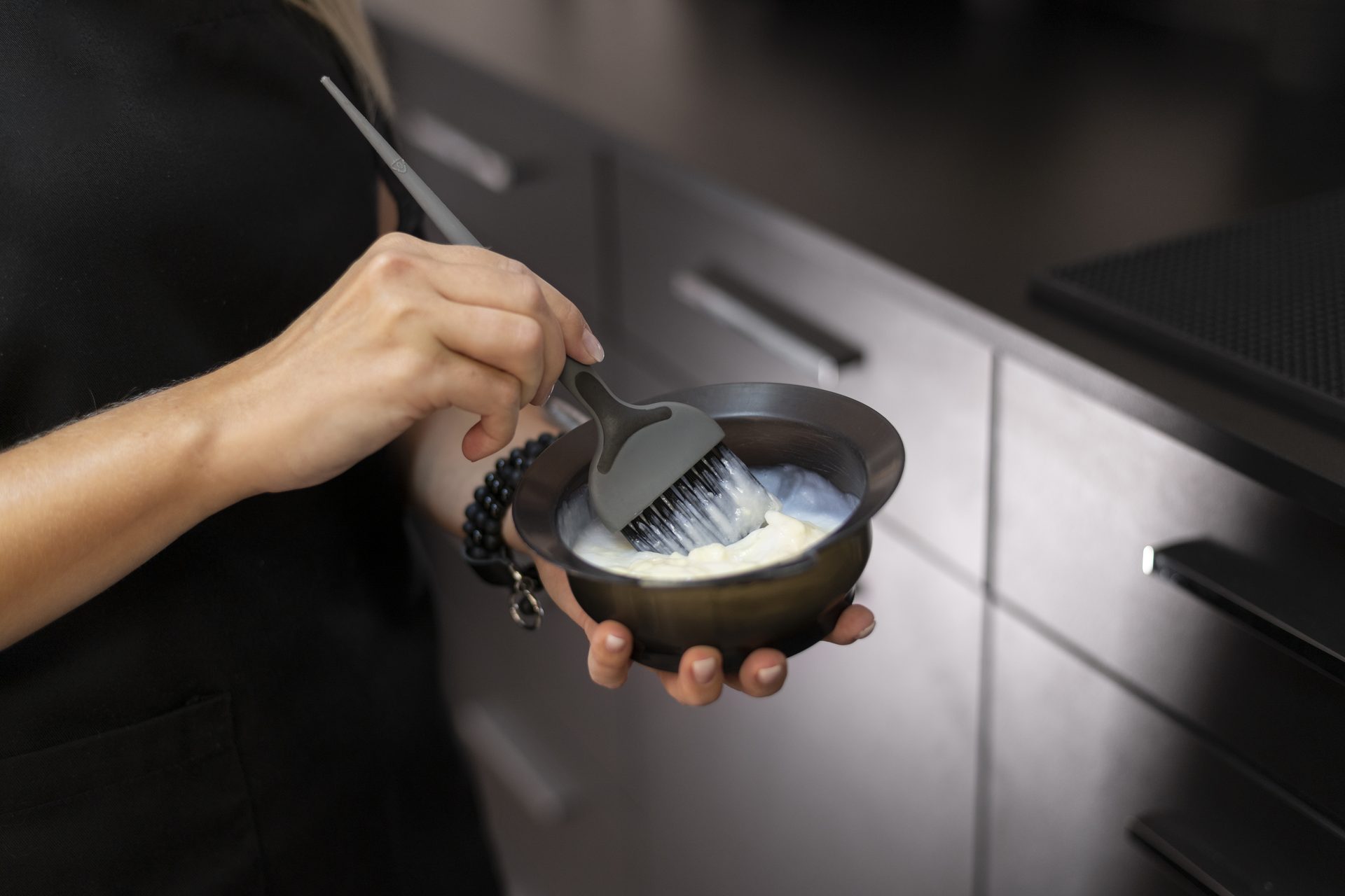 Mano mezclando crema blanca con un pincel de tinte en un bol negro junto a muebles grises
