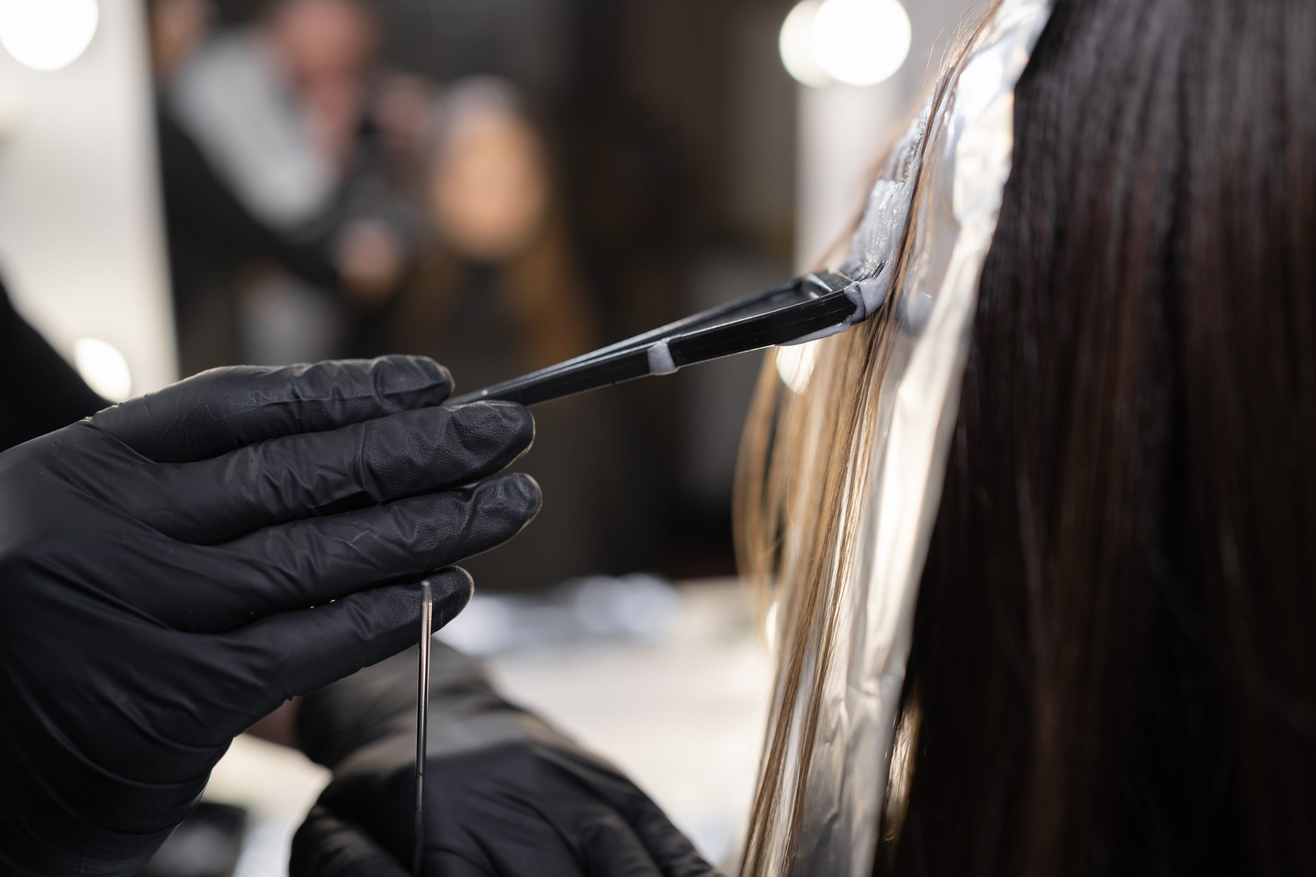 Black-gloved hands brushing product onto long dark hair laid on foil in a salon