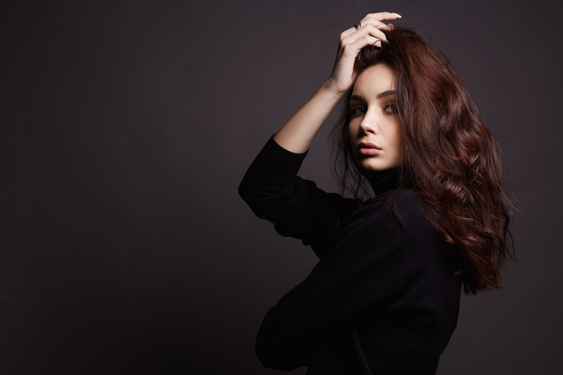 Mujer de cabello largo ondulado castaño rojizo con top negro, una mano en la cabeza, fondo oscuro