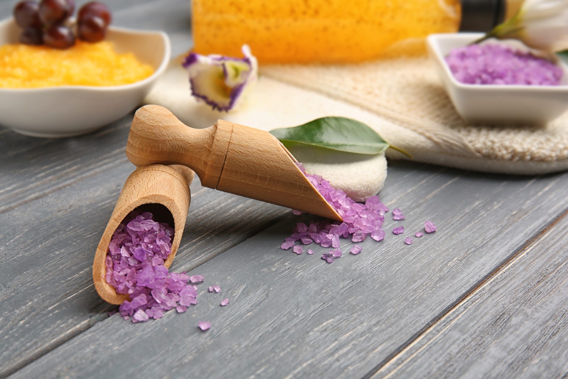 Purple bath salt spilling from a wooden scoop onto grey boards, with orange salt and grapes behind
