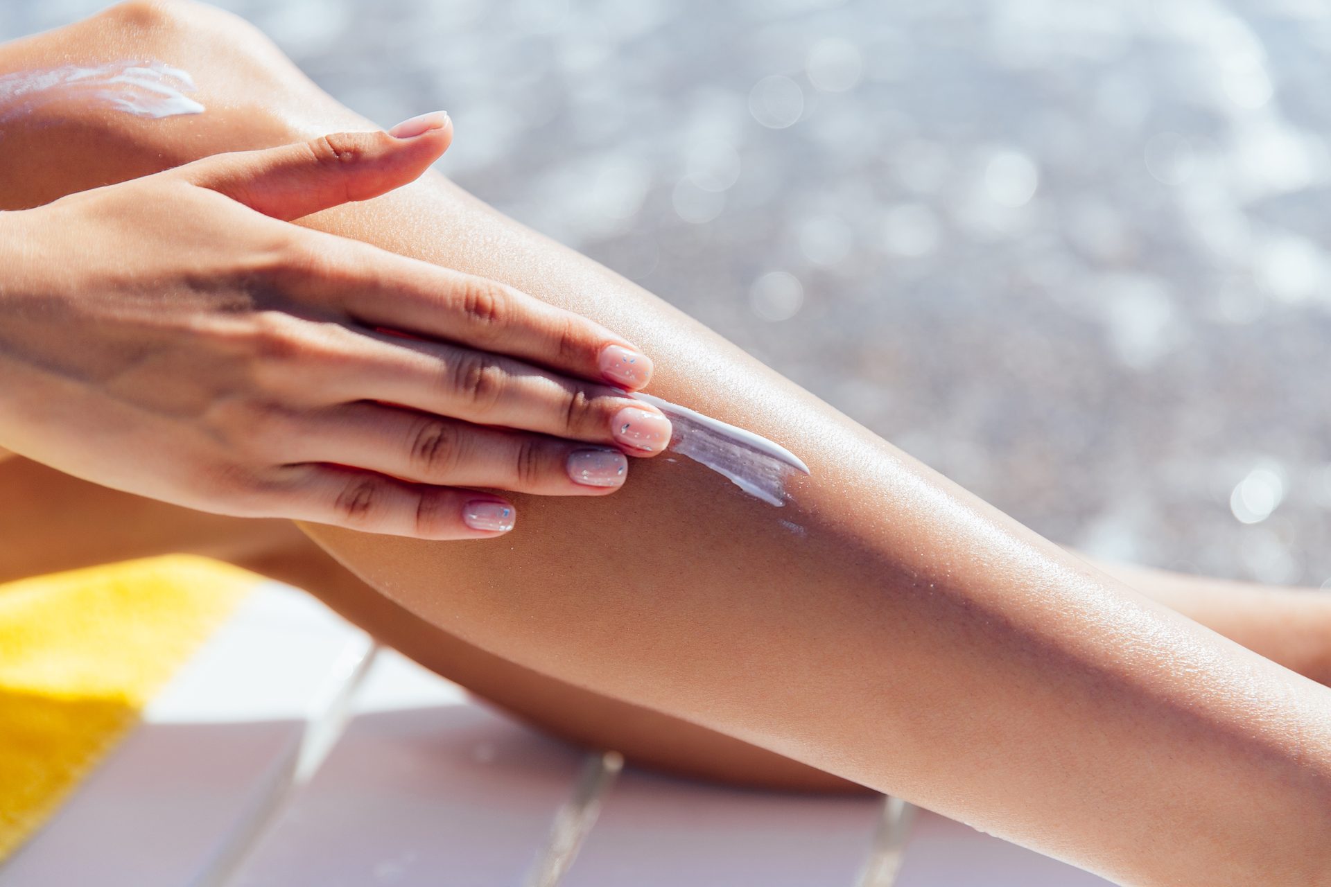Hand spreading white lotion along a tanned leg, with sparkling water behind