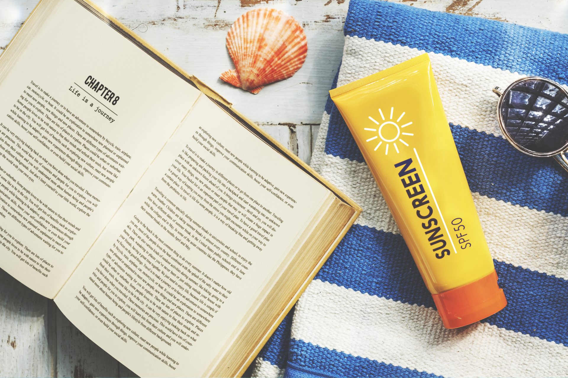 Yellow SPF 50 sunscreen tube on a blue-and-white striped towel beside an open book, shell and sunglasses