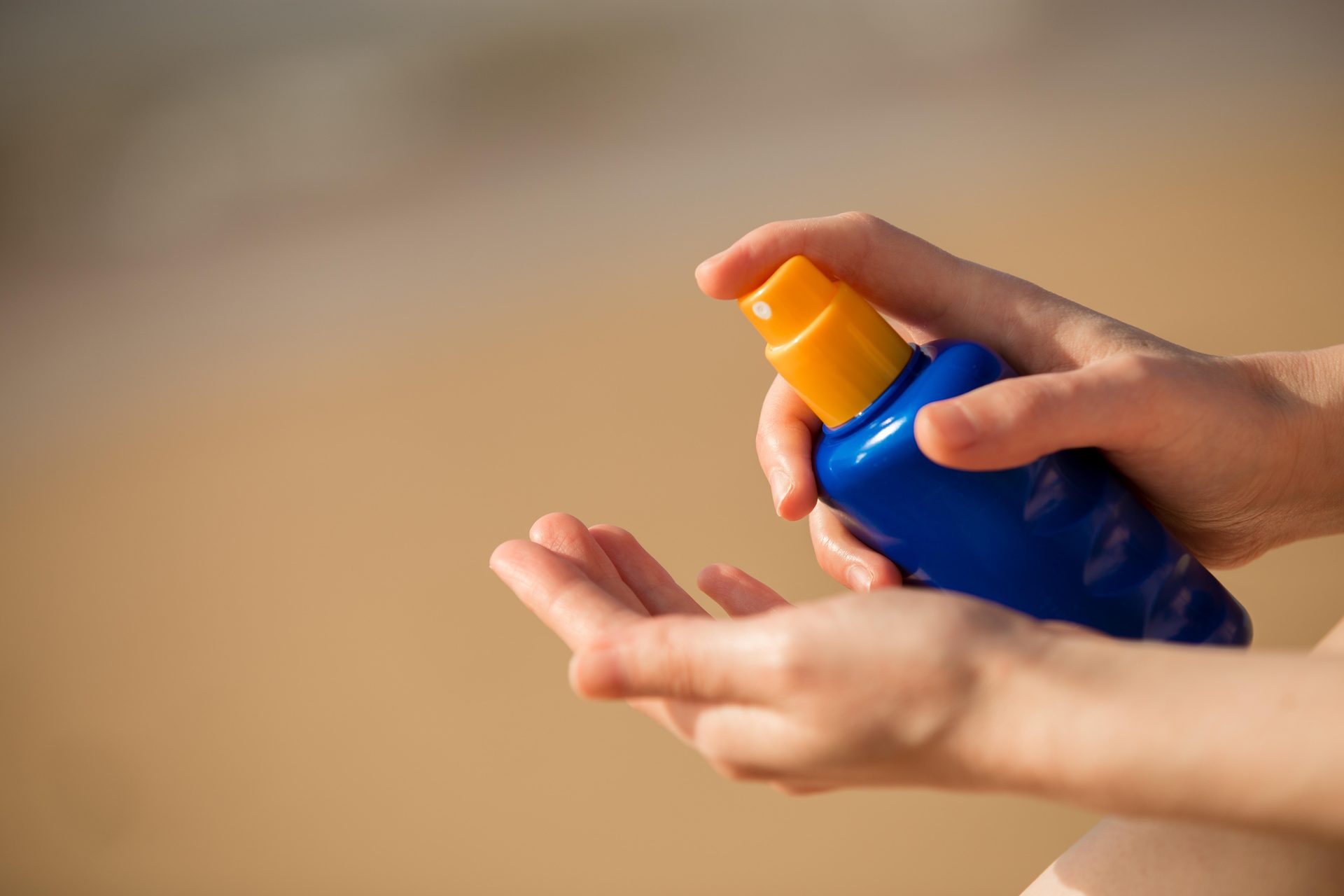 Manos sosteniendo un espray azul con tapa naranja sobre la palma abierta, playa de arena al fondo