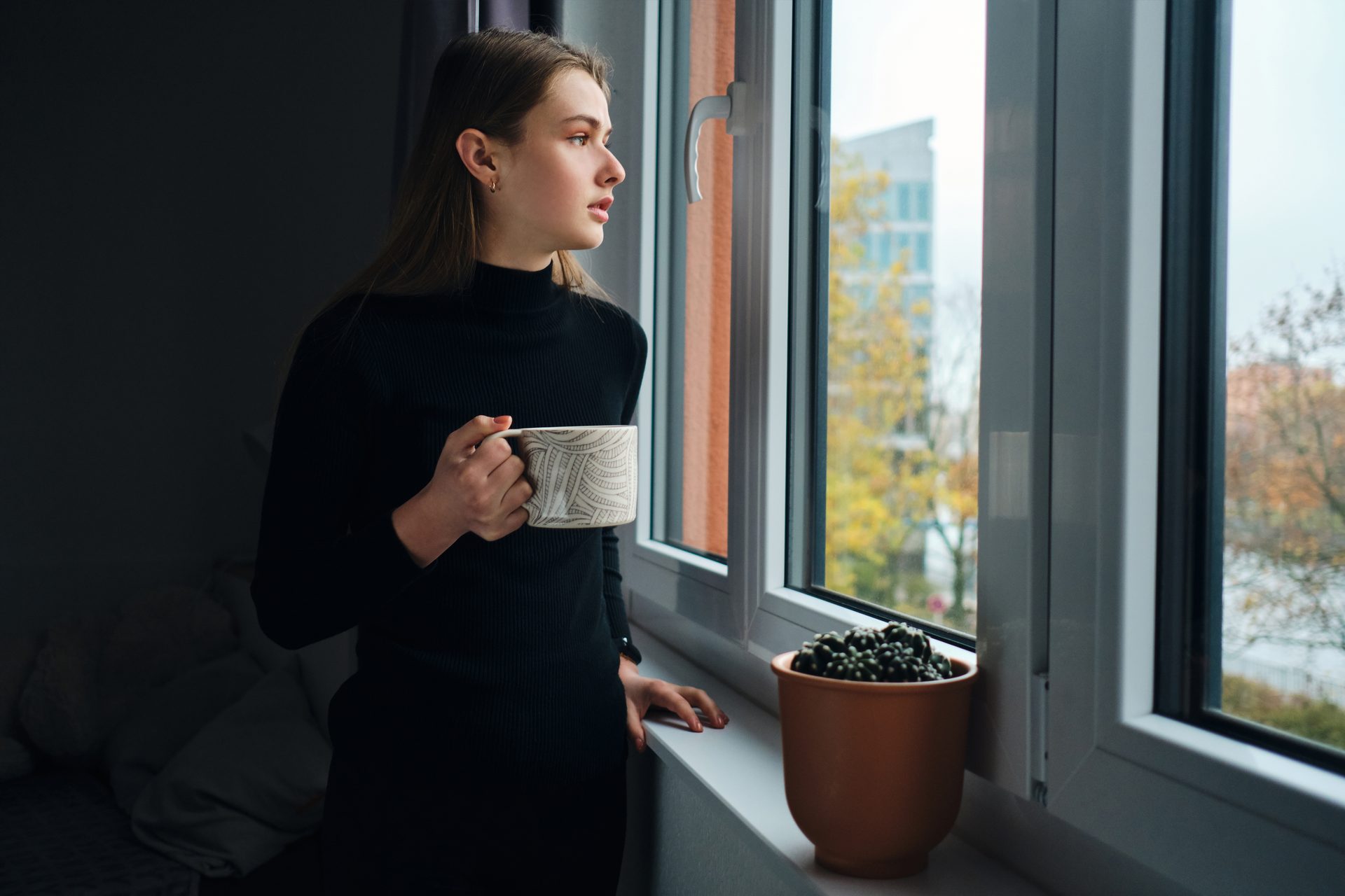 Young woman in a black jumper holding a mug, gazing out of a window on an overcast day