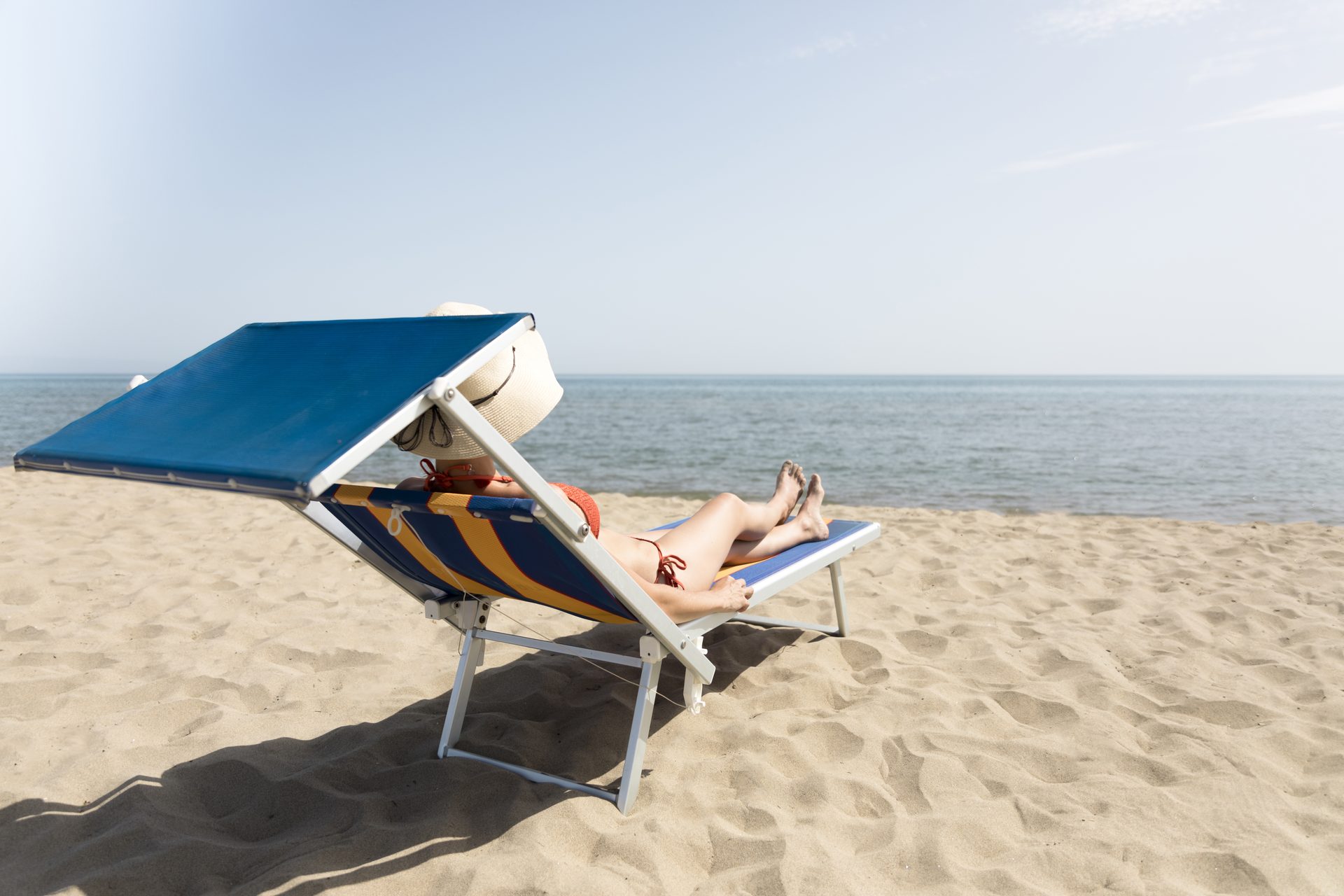 Person in a hat reclining on a striped, blue-canopied sunlounger on a sandy beach by the sea
