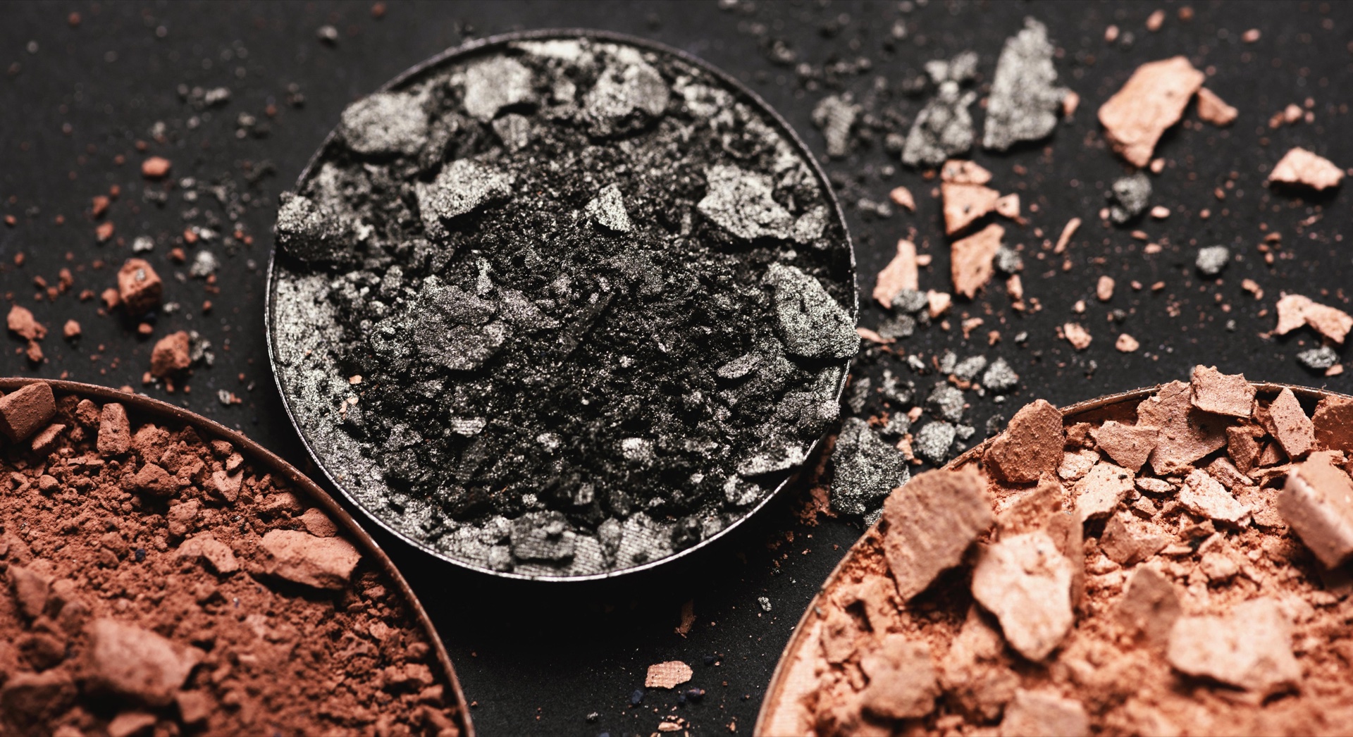 Crushed black, reddish-brown and tan pressed powders in round metal pans on a dark surface